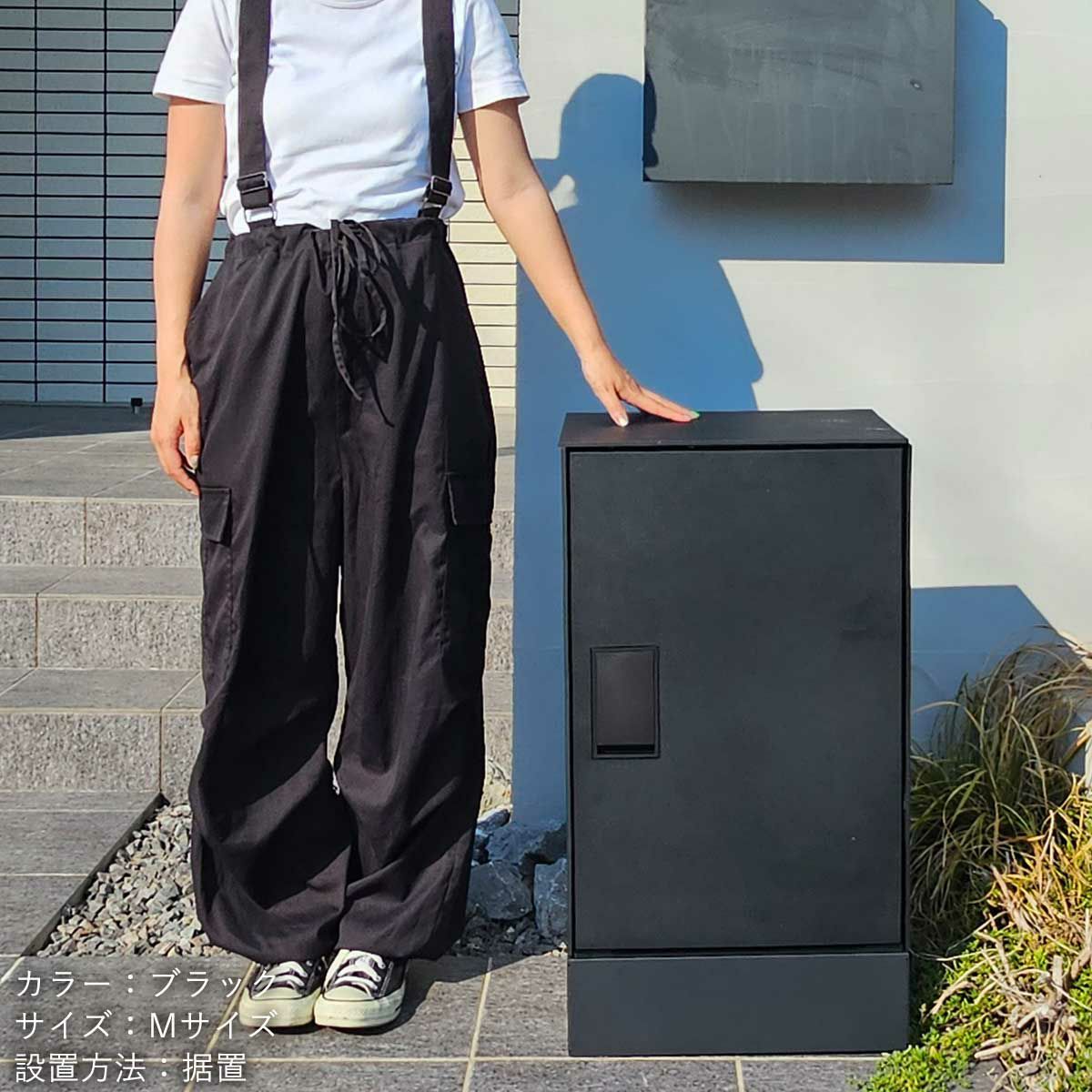 玄関扉の横に宅配ボックスが置かれていて、その横に女性が立っている設置イメージ。宅配ボックスはブラック、幅木（台座）もブラック。