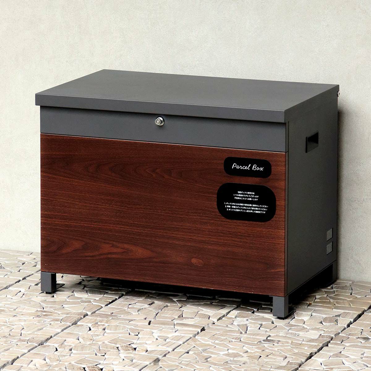 ダークグレーの宅配ボックスの設置イメージ。白い壁と石畳に置かれている。