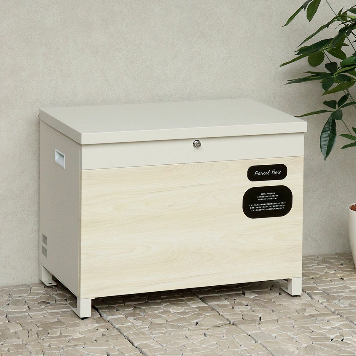 グレージュの宅配ボックスの設置イメージ。白い壁と石畳に置かれている。