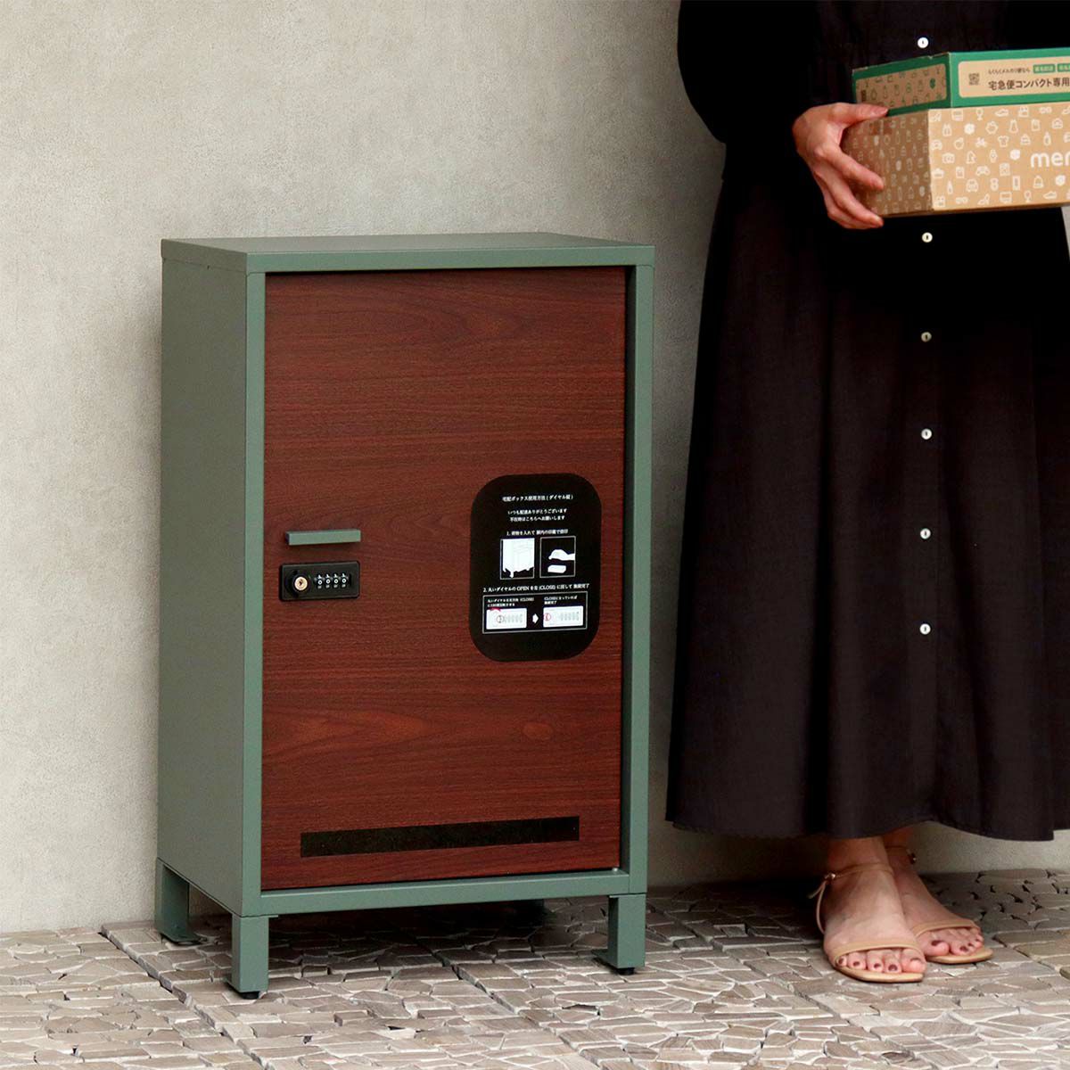 フォレストグリーンの宅配ボックスの設置イメージ。黒いワンピースを着た人が段ボールの荷物を持って側に立っている。